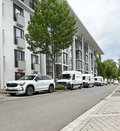 Wei&szlig;er SUV und mehrere wei&szlig;e PSM-Transporter parken vor einem modernen Wohngeb&auml;ude mit B&auml;umen entlang der Stra&szlig;e.