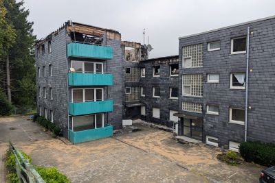 Mehrst&ouml;ckiges Wohngeb&auml;ude mit dunkler Schieferfassade und t&uuml;rkisen Balkonverkleidungen, das Dach zeigt deutliche Brandsch&auml;den.