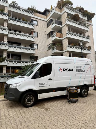 Wei&szlig;er PSM-Servicetransporter parkt vor einem mehrst&ouml;ckigen Wohngeb&auml;ude mit Balkonen, daneben steht ein Ger&auml;tekoffer.