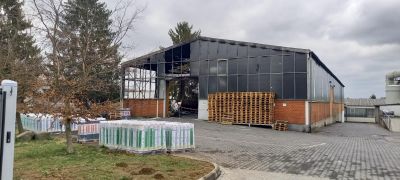 Industriehalle mit dunkler Fassade und Ziegelsockel, davor gestapelte Paletten mit verpackter Ware und ein gepflasterter Hofbereich.