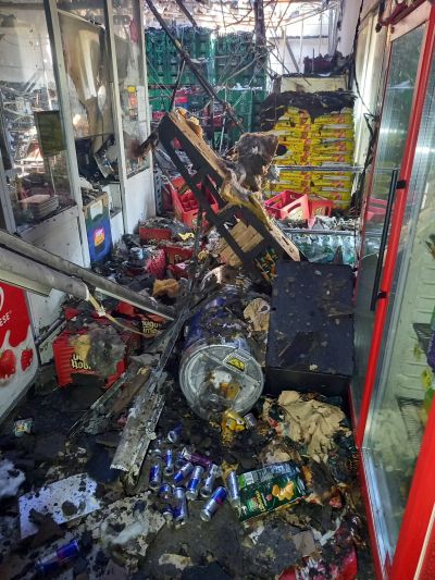 Brandschaden in einem Gesch&auml;ft mit umgest&uuml;rzten Regalen, verstreuten Dosen und ru&szlig;geschw&auml;rzten Oberfl&auml;chen.