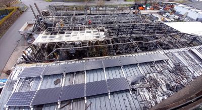 Industriegeb&auml;ude mit Solarpaneelen auf dem Dach, dahinter ein durch Brand zerst&ouml;rtes Stahlger&uuml;st ohne Verkleidung.
