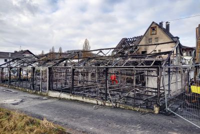 Ausgebranntes Geb&auml;ude mit verkohltem Stahlger&uuml;st, im Hintergrund ein Wohnhaus mit teilweise zerst&ouml;rtem Dach.