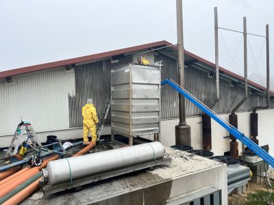 Arbeiter in gelber Schutzkleidung auf einem Industriedach neben einem Metalltank, umgeben von Schl&auml;uchen und technischen Anlagen.