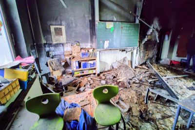 Fire-damaged classroom mit verkohlten W&auml;nden, zerst&ouml;rtem Mobiliar und Tr&uuml;mmern; gr&uuml;ne St&uuml;hle im Vordergrund.