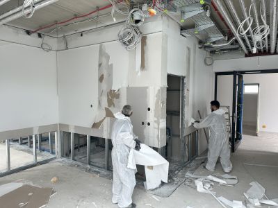 Zwei Arbeiter in Schutzanz&uuml;gen entfernen Gipskartonplatten in einem Rohbau mit sichtbaren Kabeln und Installationen.