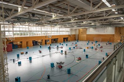 Gro&szlig;e Sporthalle mit zahlreichen blauen Boxen und Kabeln auf dem Boden, vermutlich w&auml;hrend technischer Installationsarbeiten.