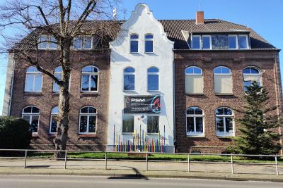 Historisches Backsteingeb&auml;ude mit wei&szlig;em Zwischengiebel und Bogenfenstern, davor ein PSM-Werbebanner und kahle B&auml;ume.