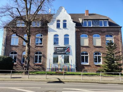 Historisches Backsteingeb&auml;ude mit wei&szlig;em Zwischengiebel und Bogenfenstern, davor ein PSM-Werbebanner und kahle B&auml;ume.