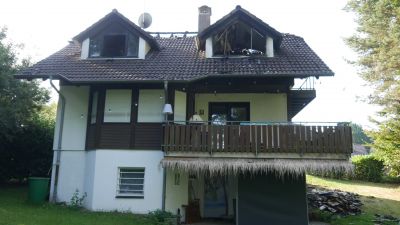 Zweist&ouml;ckiges Wohnhaus mit Balkon und dunklem Dach, das Spuren eines Brandschadens am Dachgeschoss aufweist.