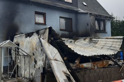 Brandschaden an einem Wohnhaus mit eingest&uuml;rztem Anbau und verkohltem Dach, abgesperrt mit rot-wei&szlig;em Flatterband.