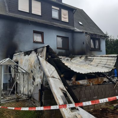 Brandschaden an einem Wohnhaus mit eingest&uuml;rztem Anbau und verkohltem Dach, abgesperrt mit rot-wei&szlig;em Flatterband.