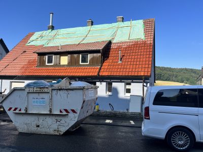 Haus mit teilweise abgedecktem Ziegeldach und Dachfenstern unter Reparatur, davor ein Container und ein wei&szlig;es Auto.