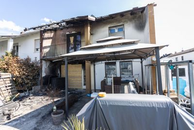 Brandschaden an einem Wohnhaus mit ru&szlig;geschw&auml;rzter Fassade, im Vordergrund Terrasse mit Sonnenschirm und M&ouml;beln.