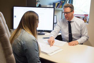 Berater und Kundin besprechen Unterlagen an einem Schreibtisch, im Hintergrund ein Computerbildschirm.