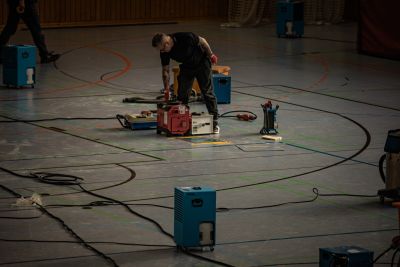 Techniker beugt sich &uuml;ber ein Ger&auml;t zur Bautrocknung in einer Sporthalle, umgeben von Kabeln und Trocknungsger&auml;ten.