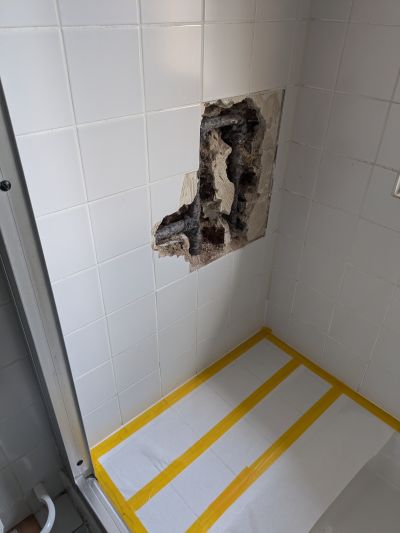 Besch&auml;digte Wandfliesen mit sichtbarem Schimmel und freigelegtem Mauerwerk, Boden mit gelbem Klebeband markiert.