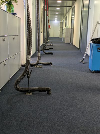 B&uuml;roflur mit mehreren Trocknungsschl&auml;uchen auf dem Teppichboden, im Hintergrund ein blaues Trocknungsger&auml;t.