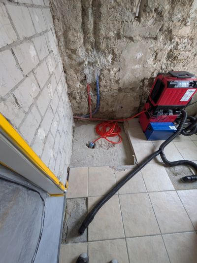 Baustelle mit freigelegten W&auml;nden, roten Rohrleitungen und einem roten Trocknungsger&auml;t neben schwarzen Schl&auml;uchen auf Fliesenboden.