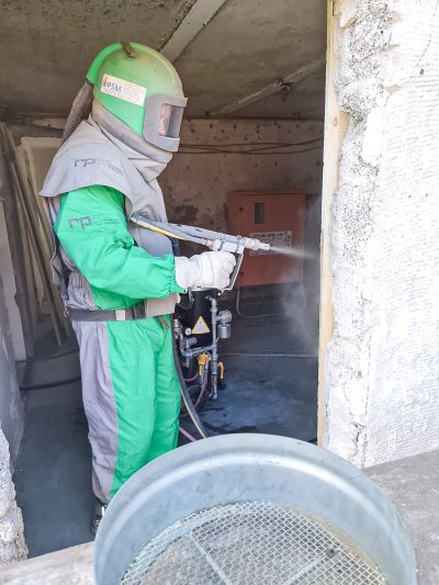 Arbeiter in Schutzausr&uuml;stung mit Helm reinigt Wand mit Hochdruckreiniger in einem Raum mit sichtbaren Brandsch&auml;den.