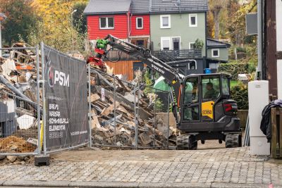 Bagger r&auml;umt Tr&uuml;mmer und Bauschutt hinter einem Bauzaun mit PSM-Werbung, im Hintergrund Wohnh&auml;user.