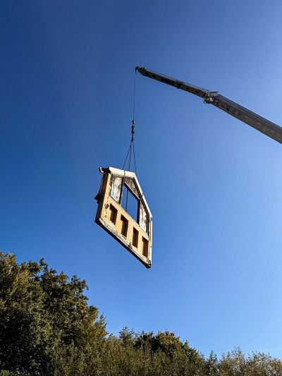 Ein Kran hebt ein besch&auml;digtes, dreieckiges Hausteil mit verkohlten Balken in den blauen Himmel &uuml;ber B&auml;umen.