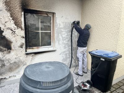 Arbeiter reinigt ru&szlig;geschw&auml;rzte Hauswand nach Brandschaden mit Schleifger&auml;t, daneben ein verru&szlig;tes Fenster.