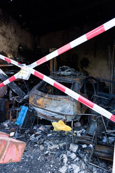 Ausgebranntes Fahrzeug in einer verru&szlig;ten Garage, abgesperrt mit rot-wei&szlig;em Absperrband, umgeben von Tr&uuml;mmern und Asche.