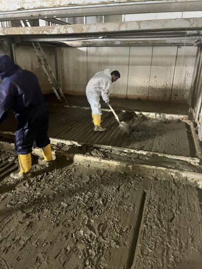 Zwei Arbeiter in Schutzkleidung und Gummistiefeln beseitigen Schlamm in einem &uuml;berfluteten Industrieraum.