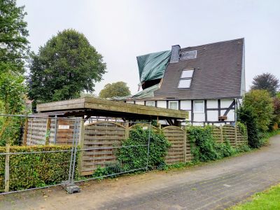 Sturmschaden am Dach eines Fachwerkhauses mit Planenabdeckung, im Vordergrund Zaun und Carport.