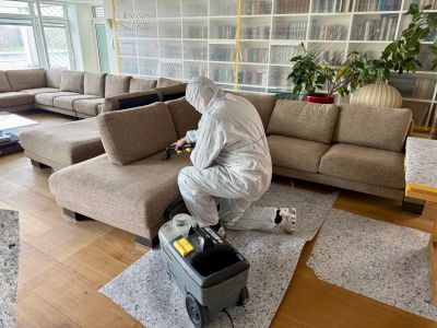 Techniker in Schutzanzug reinigt Polstersofa mit professionellem Reinigungsger&auml;t in modernem Wohnzimmer.