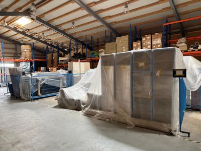 Lagerhalle mit foliertem Mobiliar und Kartons auf Palettenregalen, im Hintergrund weitere Lagerbest&auml;nde.