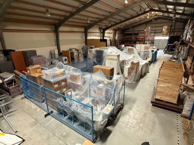 Lagerhalle mit blauen Gitterwagen voller verpackter M&ouml;bel und Kartons sowie Holzpaletten im Hintergrund.