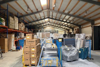 Lagerhalle mit gestapelten Kartons, verpackten M&ouml;beln und Waren auf Paletten unter einem Dach mit Metalltr&auml;gern.