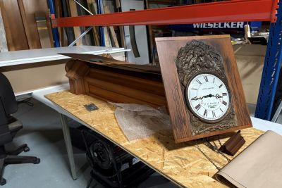 Antike Wanduhr mit ornamentiertem Holzrahmen und Ziffernblatt, daneben ein aufgeklapptes Uhrengeh&auml;use auf einer Werkbank.