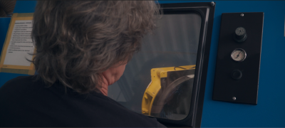 Person mit grauen Haaren blickt durch ein Sichtfenster einer Maschine, neben einem Bedienfeld mit Reglern.