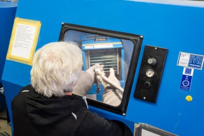 Person mit wei&szlig;en Haaren arbeitet an einer blauen Industriemaschine mit Sichtfenster, Manometern und Schaltern.
