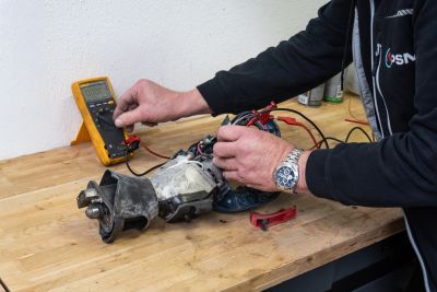Techniker pr&uuml;ft einen alten Elektromotor mit einem gelben Multimeter auf einer Werkbank.