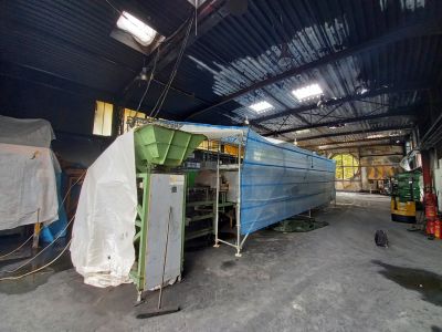 Industriehalle mit langer, blau abgedeckter Maschinenanlage auf Ger&uuml;stb&ouml;cken und gr&uuml;ner Maschine mit Plane im Vordergrund.