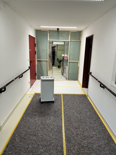 Flur mit Handl&auml;ufen, Schutzfolie an einer T&uuml;r und einem Luftreiniger auf schwarzem Teppich mit gelber Klebeband-Markierung.