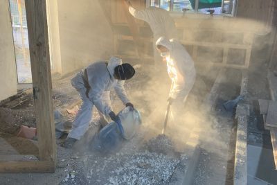 Zwei Arbeiter in Schutzanz&uuml;gen entfernen staubige Schadstoffe in einem Rohbau, umgeben von aufgewirbeltem Staub.