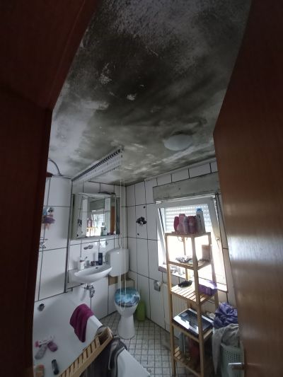 Badezimmer mit stark schimmelbefallener Decke, wei&szlig; gekachelten W&auml;nden, Waschbecken, Badewanne und Regal mit Utensilien.