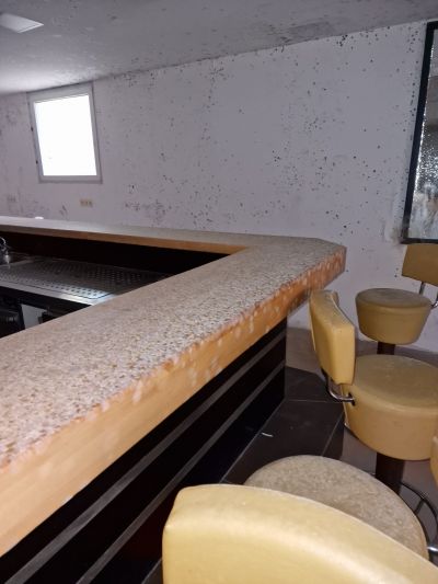 Verschimmelte Wand &uuml;ber einer Bartheke mit gelben Barhockern in einem Kellerraum.