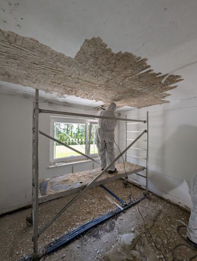 Arbeiter in Schutzanzug entfernt schimmelbefallenen Deckenverputz auf einem Ger&uuml;st in einem Innenraum.