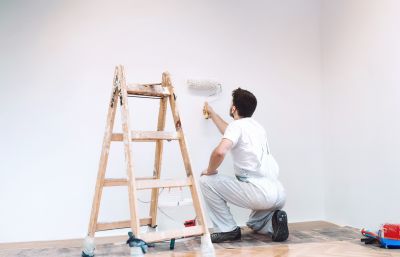 Handwerker in Arbeitskleidung streicht eine Wand mit einer Farbrolle, daneben steht eine Holzleiter.