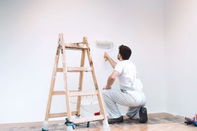 Handwerker in Arbeitskleidung streicht eine Wand mit einer Farbrolle, daneben steht eine Holzleiter.