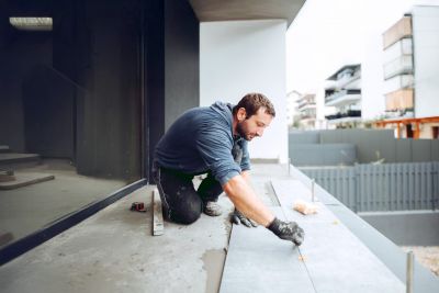 Handwerker verlegt Bodenplatten auf einer Terrasse, im Hintergrund Geb&auml;ude und ein Zaun.