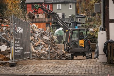 Bagger r&auml;umt Bauschutt hinter einem Zaun mit PSM-Werbung, im Hintergrund Wohnh&auml;user.