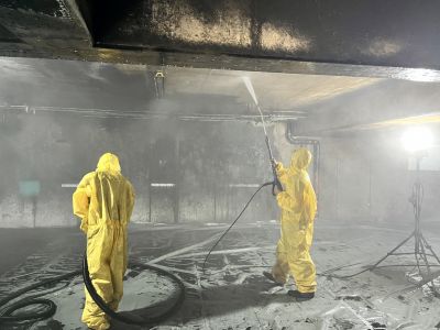 Zwei Arbeiter in gelben Schutzanz&uuml;gen reinigen eine verru&szlig;te Decke mit Hochdruckreiniger in einem Industrieraum.