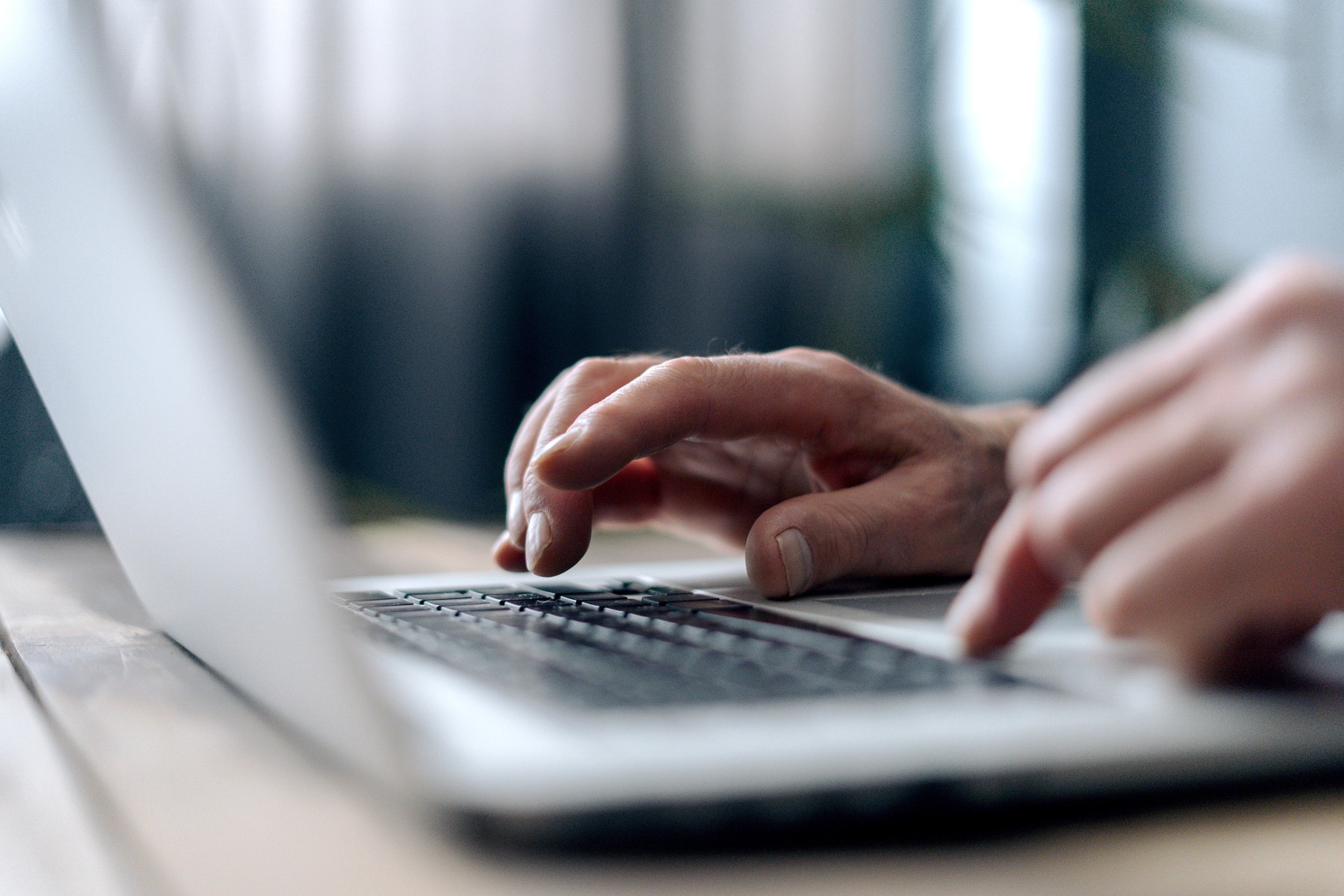 Nahaufnahme von Händen, die auf einer Laptop-Tastatur tippen, im Hintergrund unscharfe Pflanzen und Fenster.